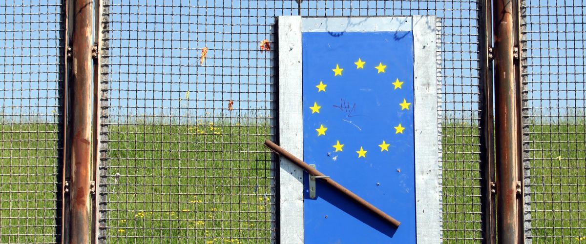 Maschendrahtzaun mit verriegelter Tür mit EU-Symbol auf der grünen Wiese.